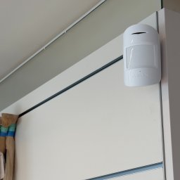 Defence Sp. z o. o. - Biała czujka ruchu Ajax i kamera Dahua zamontowane na ścianie. Widok z dołu, oświetlenie dzienne. Elementy systemu alarmowego wewnątrz pomieszczenia.