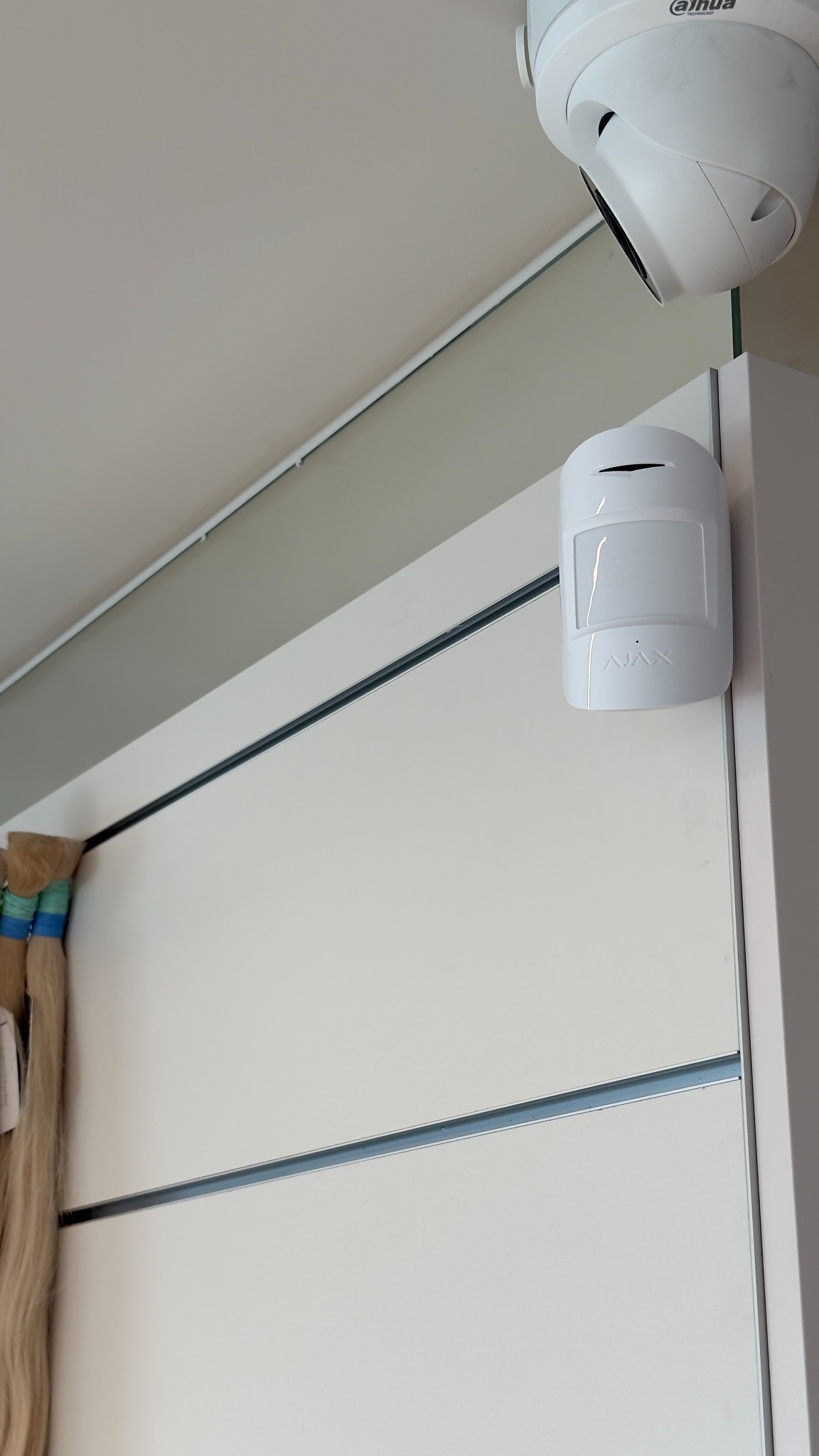 Biała czujka ruchu Ajax i kamera Dahua zamontowane na ścianie. Widok z dołu, oświetlenie dzienne. Elementy systemu alarmowego wewnątrz pomieszczenia.
