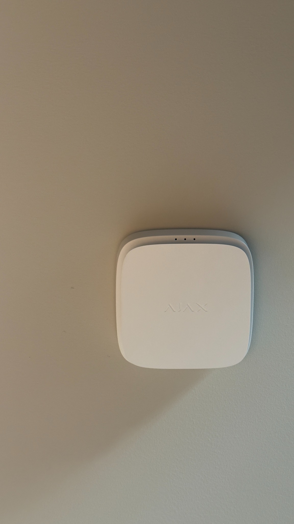 Zamontowany na ścianie, biały, bezprzewodowy czujnik dymu, CO i gwałtownego wzrostu temperatury Ajax 3 w 1. Widoczne subtelne logo producenta.