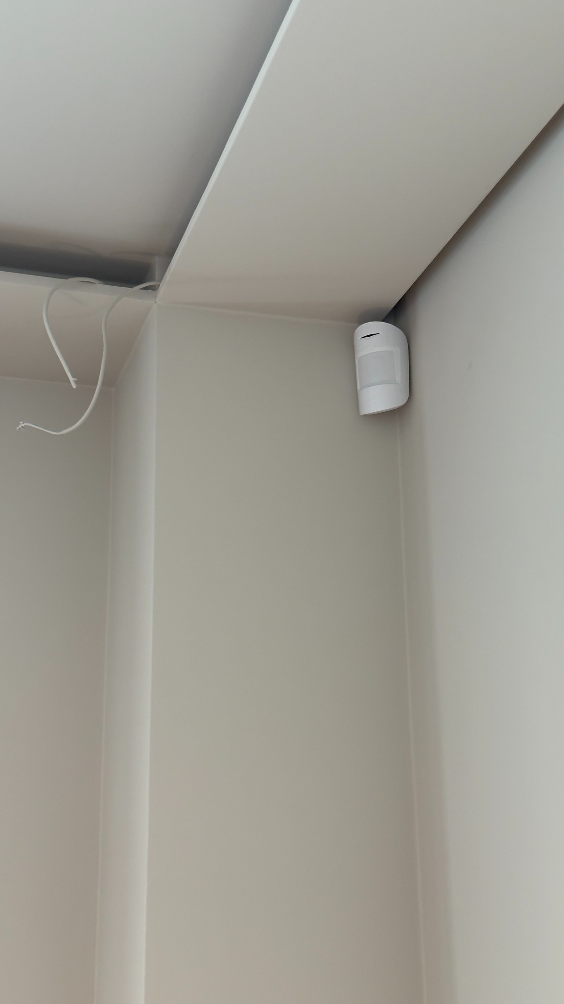 Zamontowana, bezprzewodowa czujka ruchu i rozbicia szyby z podwójną weryfikacją, umieszczona w rogu pomieszczenia o jasnych ścianach i suficie.