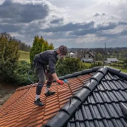Pracownik w masce maluje dach&oacute;wkę natryskowo na czarno, kontrastując z pomarańczową, niemalowaną częścią. W tle zabudowa wiejska i pochmurne niebo.