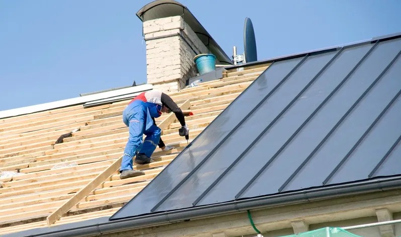 Dekarz w niebieskim kombinezonie montuje metalowe pokrycie dachu na drewnianej konstrukcji, widoczny komin i rynna, jasne niebo.