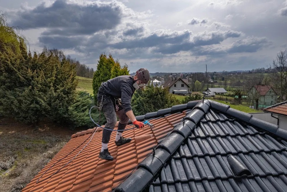 Pracownik w masce maluje dachówkę natryskowo na czarno, kontrastując z pomarańczową, niemalowaną częścią. W tle zabudowa wiejska i pochmurne niebo.