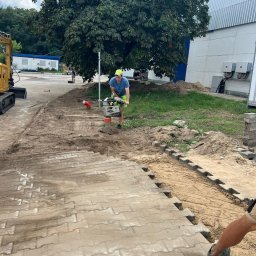 Usługi koparką DK - Pracownik ubija piasek zagęszczarką skoczkową przed ułożeniem kostki brukowej. Częściowo ułożona kostka, piasek, maszyna budowlana, drzewo w tle.