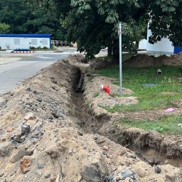 Usługi koparką DK - Wykopany rów z rurami na tle zieleni i budynku. Widoczne ślady ziemi i kamieni. Rów przygotowany pod instalacje, z elementami przyłączeniowymi.