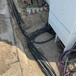 Usługi koparką DK - Kable ułożone w wykopie, prowadzące do białej skrzynki HAK. Widoczna stalowa listwa, cegły w tle. Instalacja kabli w ziemi, widok z góry.