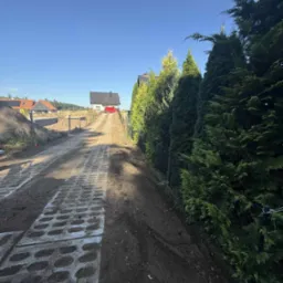 Podjazd z ażurowych płyt betonowych w trakcie budowy, z widokiem na dom z czerwonym samochodem. Po prawej stronie rząd tuj.