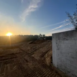 Niwelacja terenu pod budowę: teren wyrównany, widoczne ślady opon, w tle las i koparka, w prawym dolnym rogu fragment betonowego muru, zachód słońca.