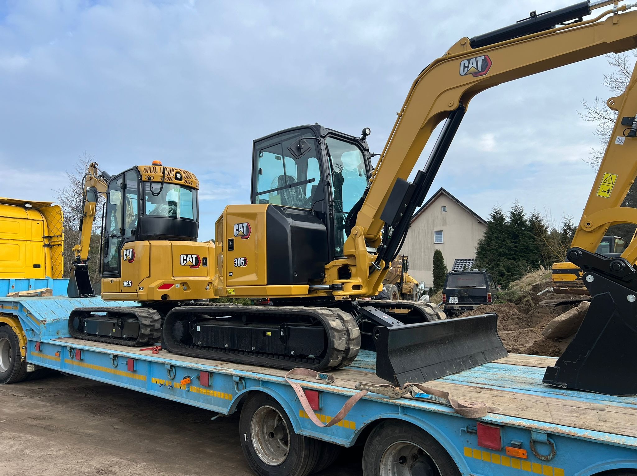 Żółty mini-koparka CAT 305 na niebieskiej platformie transportowej, widoczny fragment kabiny i ramię z łyżką, w tle dom jednorodzinny i inne maszyny budowlane.