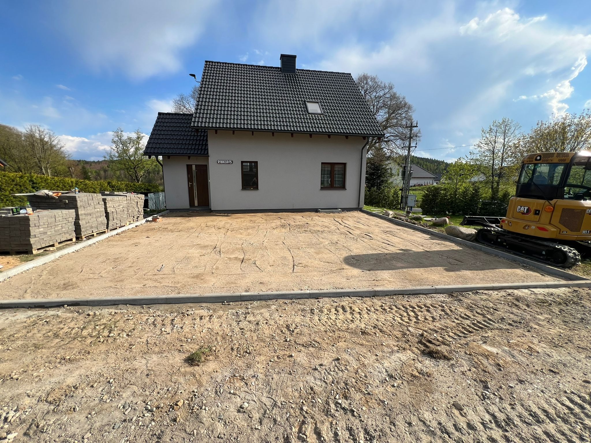 Przygotowany teren pod układanie kostki brukowej przed domem. Widoczny mini-koparka, paleta z kostką, obrzeża betonowe i wyrównany piasek. Słoneczny dzień.