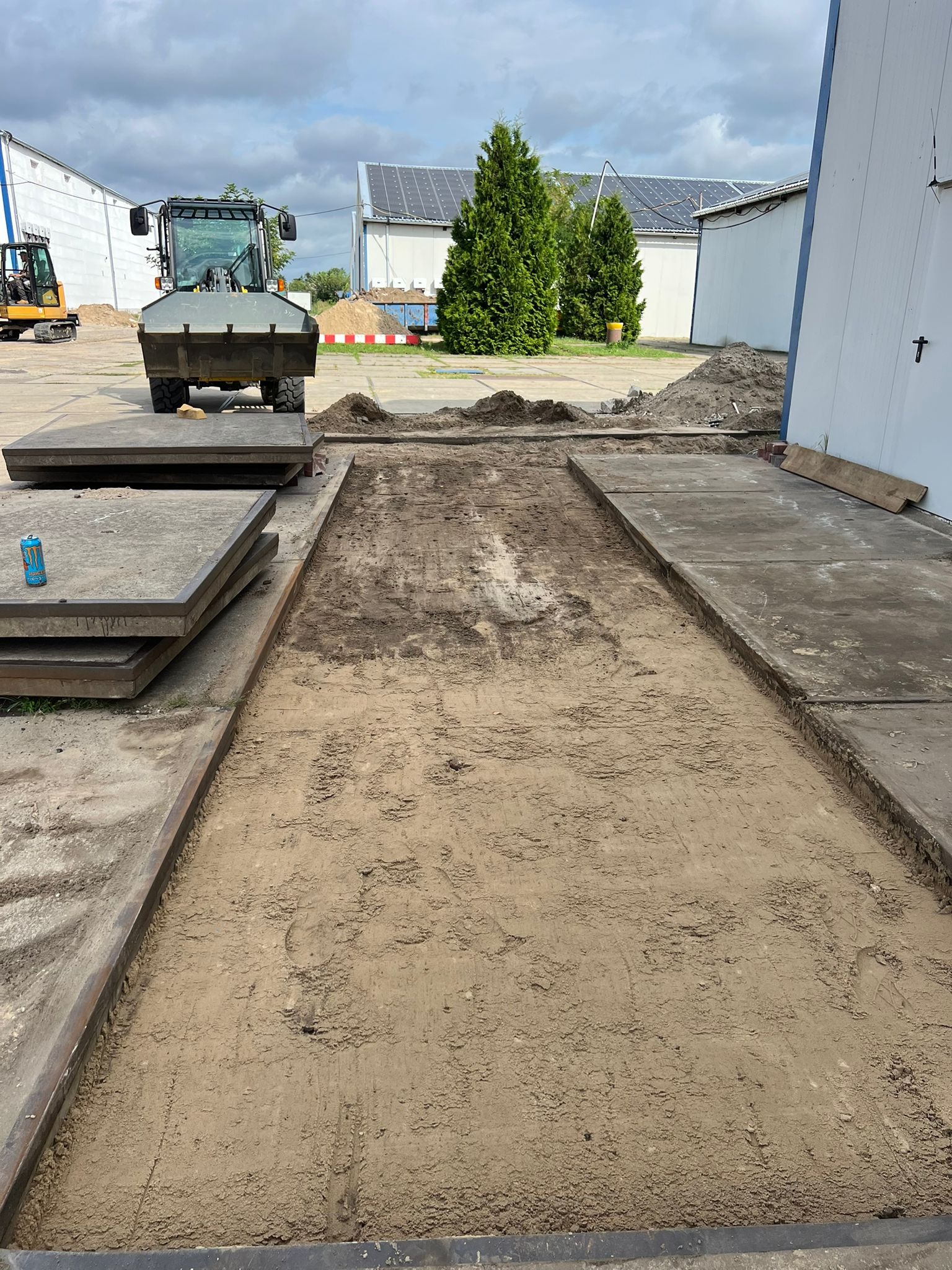 Wyrównany teren pod budowę z użyciem ciężkiego sprzętu, widoczne płyty betonowe i mała koparka w tle. Tło stanowią budynki z panelami słonecznymi i drzewa.