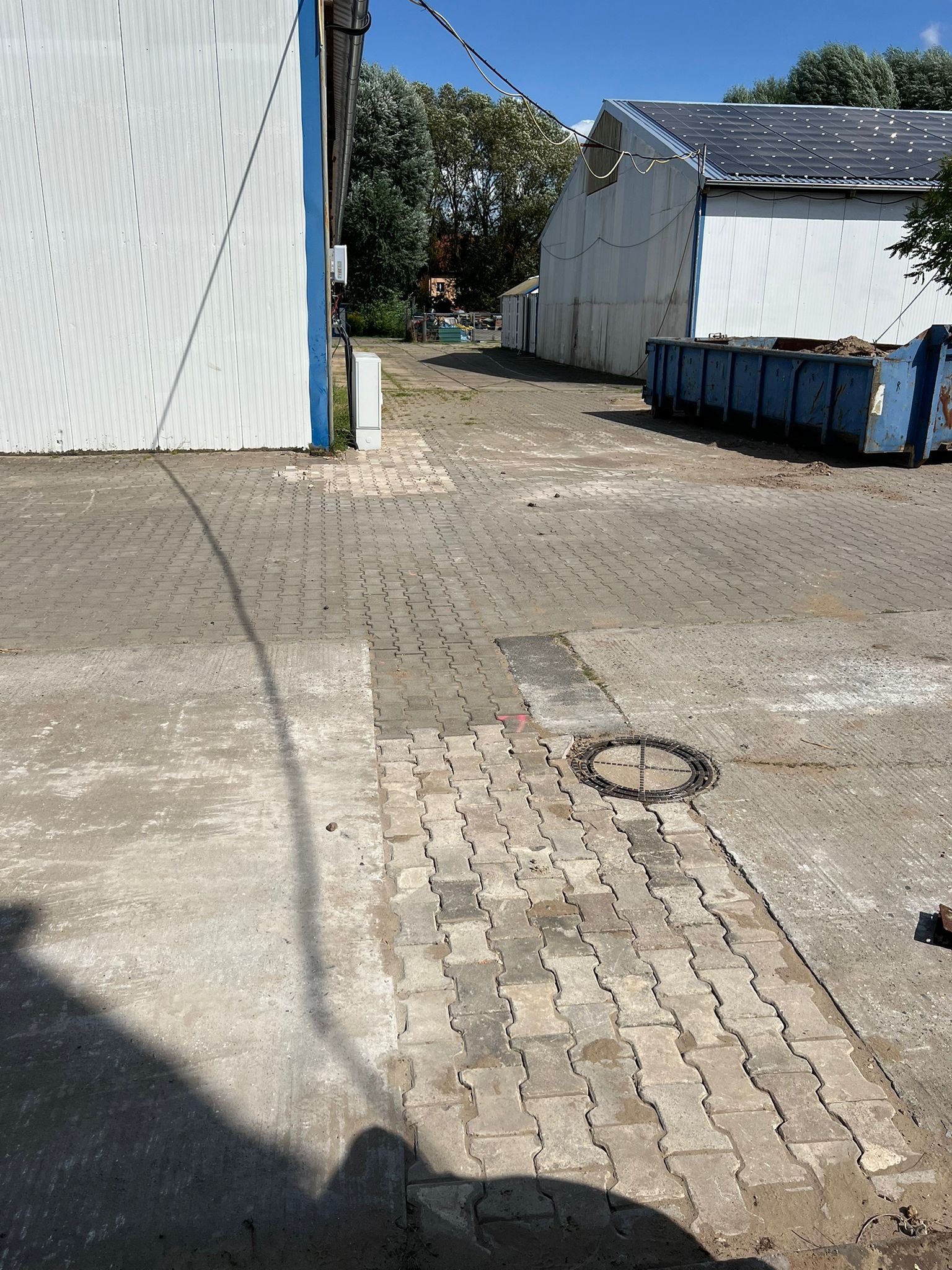 Ułożona kostka brukowa na utwardzonym placu z budynkami w tle. Widoczny fragment z kostką oraz studzienka kanalizacyjna. Obok stoi kontener.