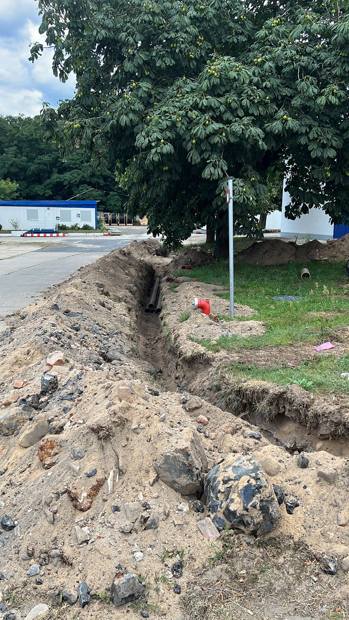 Wykopany rów z rurami na tle zieleni i budynku. Widoczne ślady ziemi i kamieni. Rów przygotowany pod instalacje, z elementami przyłączeniowymi.
