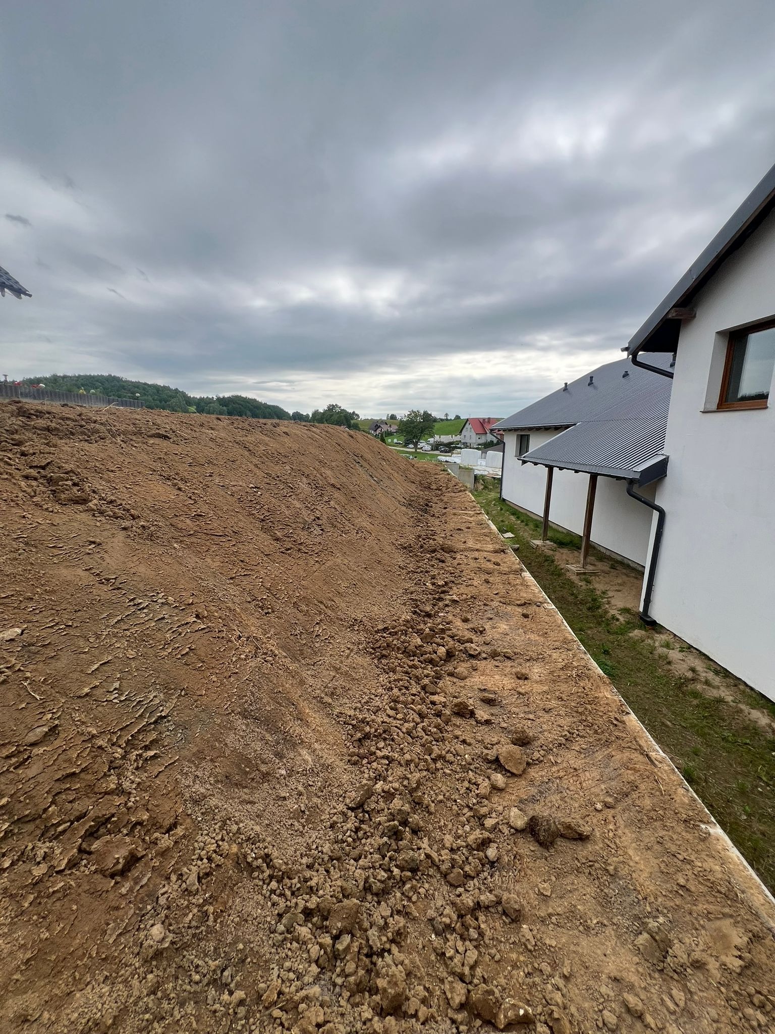 Wyrównany teren przy nowym budynku w pochmurny dzień. Widoczna świeża ziemia, dom z szarą dachówką i fragment sąsiedniej zabudowy w tle.