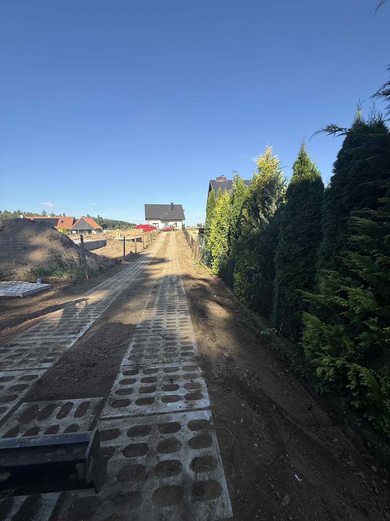 Fragment układania ażurowej kostki brukowej na podjeździe. W tle dom jednorodzinny i zielone tuje. Widoczne nierówności terenu i przygotowane podłoże.