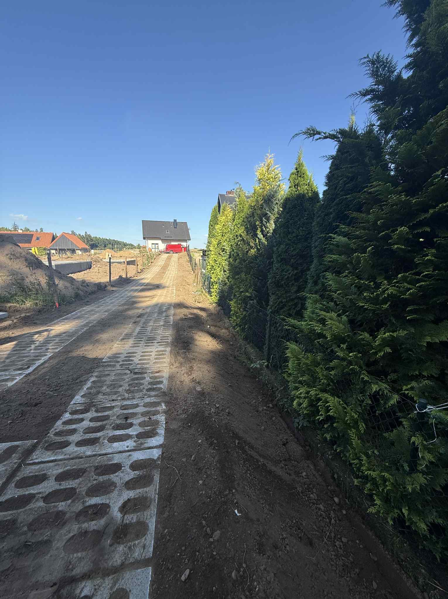 Podjazd z ażurowych płyt betonowych w trakcie budowy, z widokiem na dom z czerwonym samochodem. Po prawej stronie rząd tuj.