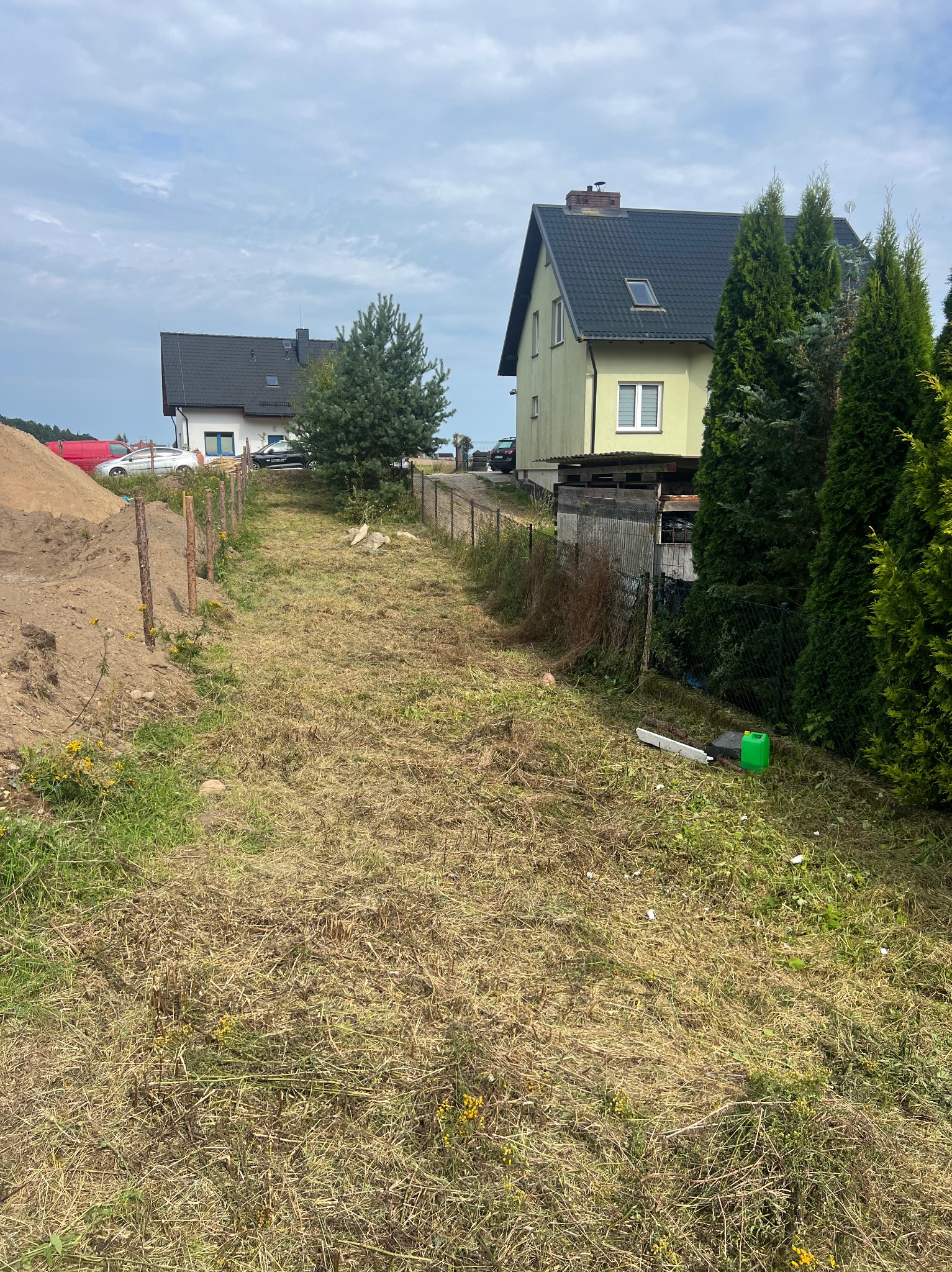 Działka z wytyczonym terenem pod budowę, widoczne domy w tle. Słupki ogrodzeniowe wzdłuż ścieżki, hałda ziemi po lewej stronie kadru.