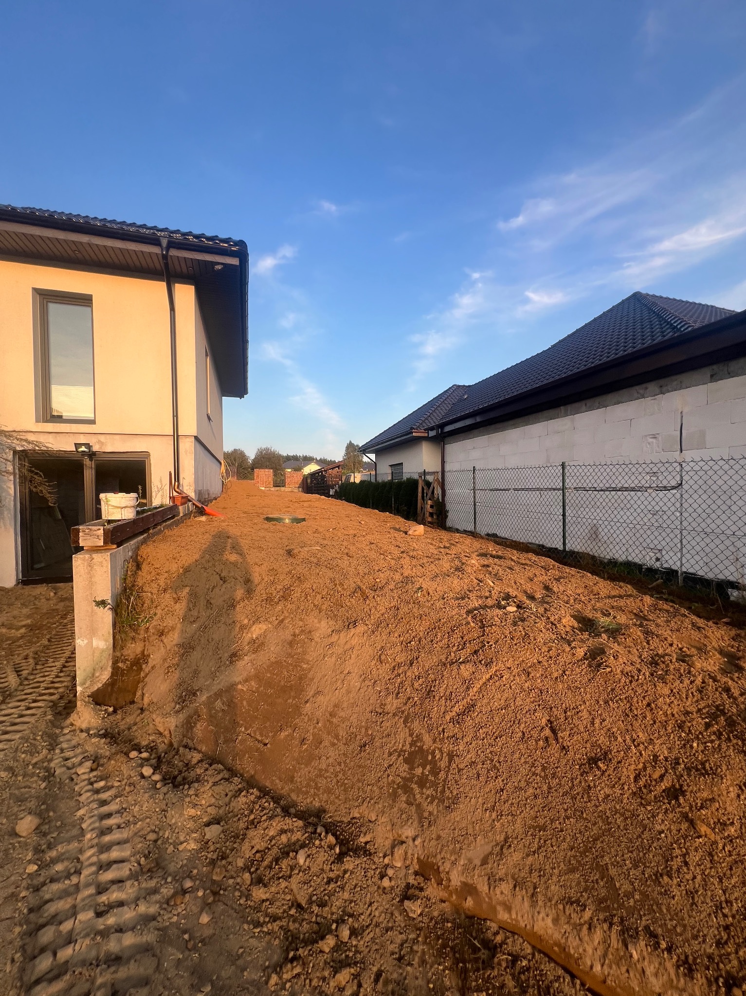 Wyrównany teren pod budowę domu jednorodzinnego, widoczne ślady opon, częściowo ogrodzony teren, błękitne niebo. W tle zabudowa jednorodzinna.