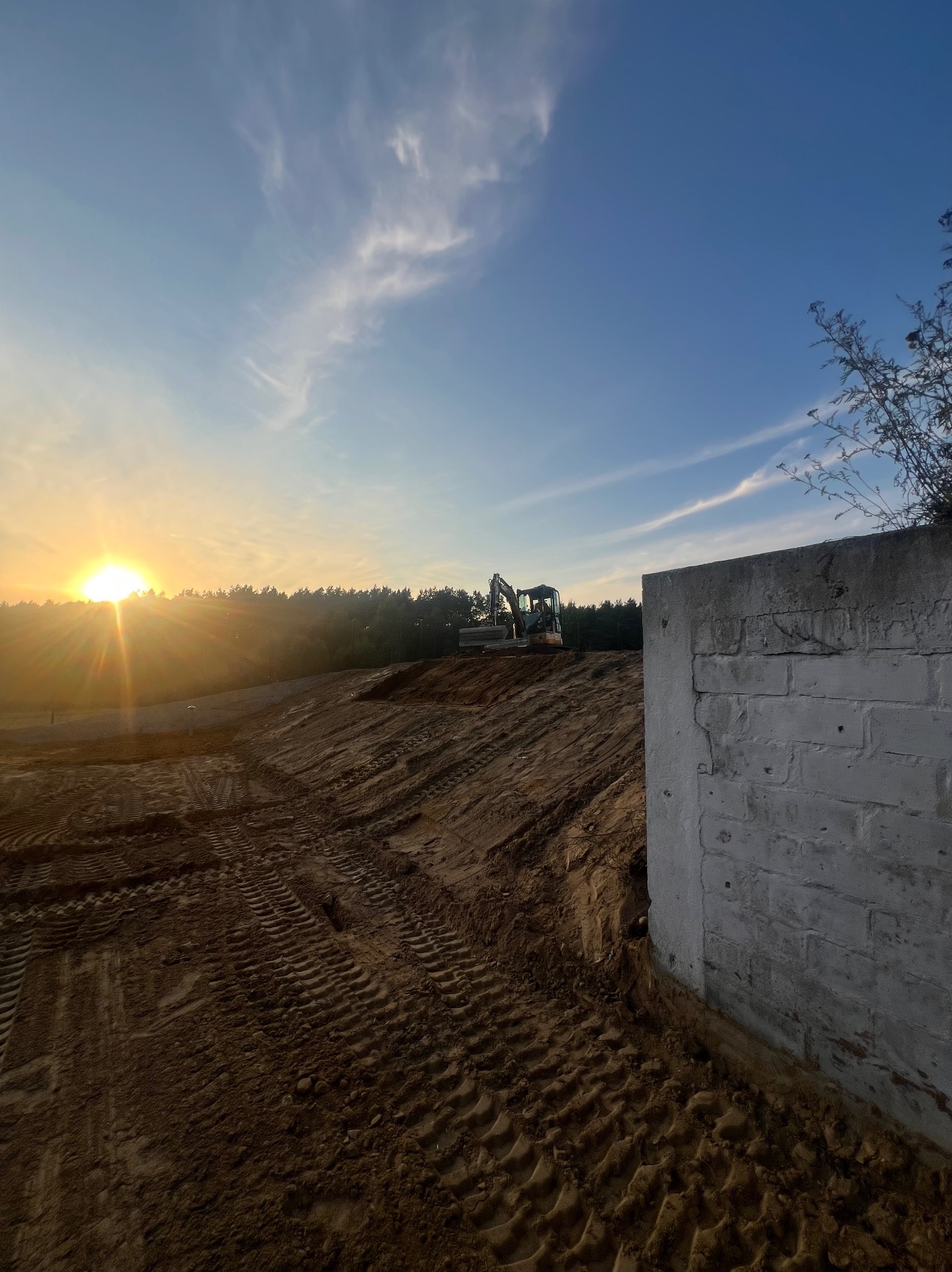 Niwelacja terenu pod budowę: teren wyrównany, widoczne ślady opon, w tle las i koparka, w prawym dolnym rogu fragment betonowego muru, zachód słońca.