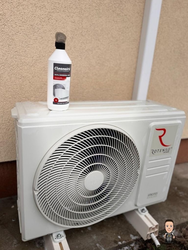 Biała jednostka zewnętrzna klimatyzacji Rotenso z butelką środka czyszczącego Cleanairix na górze, ustawiona na betonowym podłożu przy beżowej ścianie budynku.