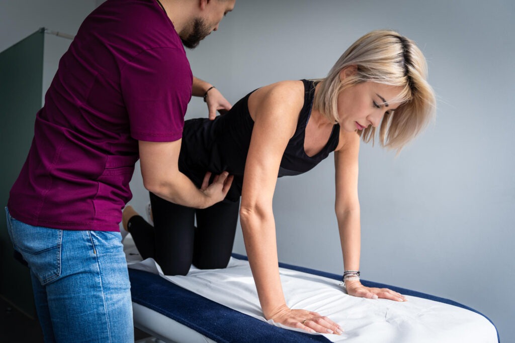 Kobieta w pozycji na czworakach poddawana badaniu palpacyjnemu pleców przez mężczyznę w fioletowej koszulce na kozetce w gabinecie.