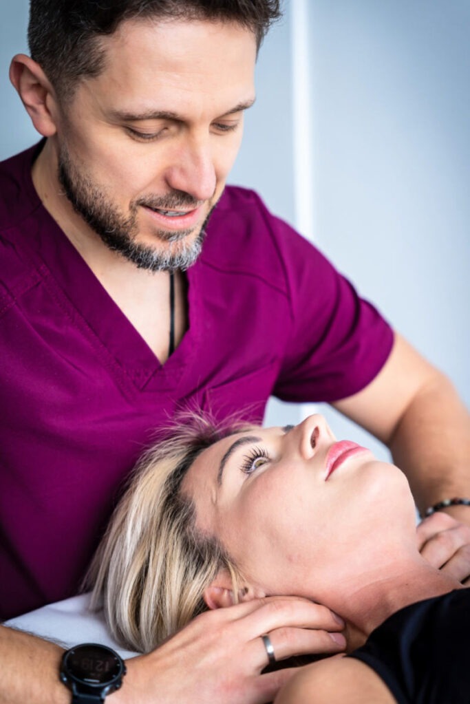 Sesja manualna: terapeuta w bordowej koszuli delikatnie masuje szyję pacjentki leżącej na plecach. Widoczny zegarek na nadgarstku terapeuty.