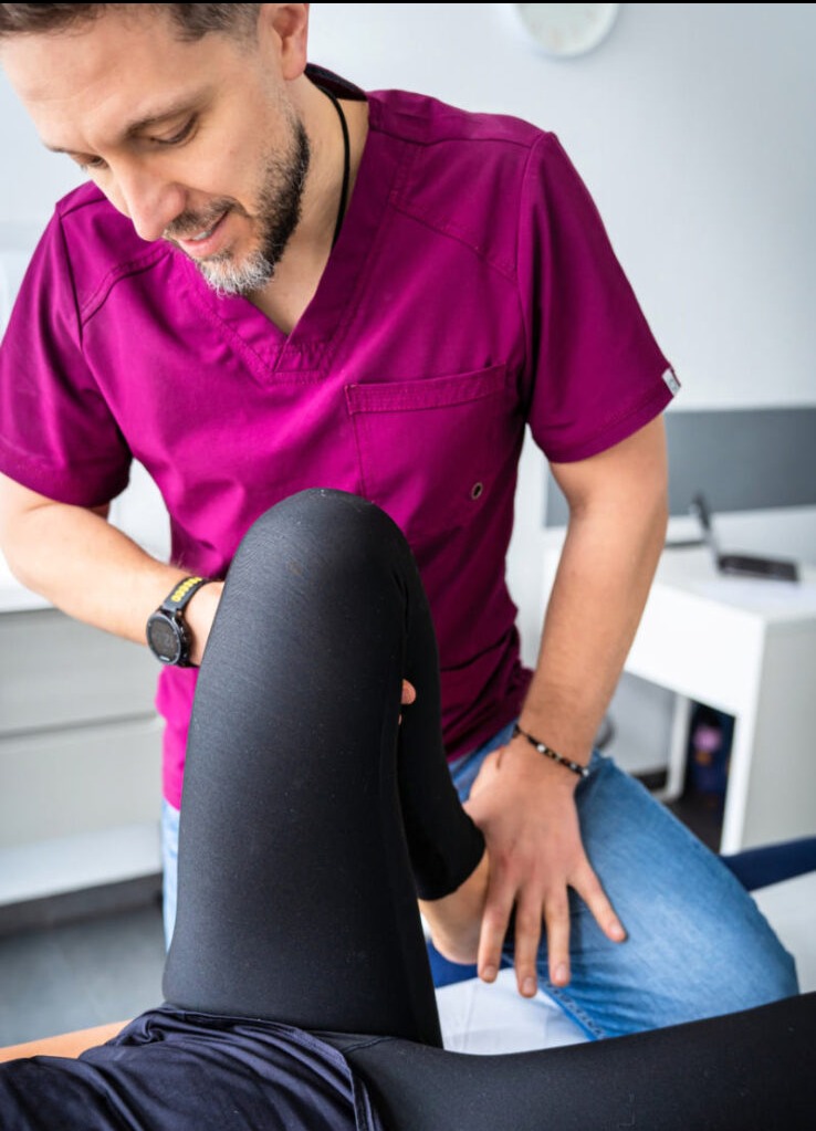 Osoba w fuksjowej bluzie uciskająca kolano pacjenta leżącego na leżance w gabinecie. Na nadgarstku zegarek sportowy.
