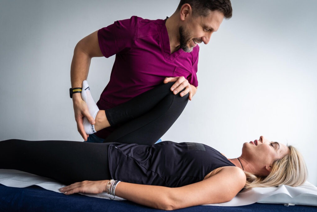 Mężczyzna w bordowej bluzie rozciąga nogę leżącej pacjentki na niebieskim łóżku rehabilitacyjnym. Uśmiechnięty fizjoterapeuta prowadzi sesję rozciągania.