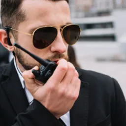 Mężczyzna w garniturze i okularach przeciwsłonecznych rozmawia przez krótkofalówkę. W tle budynek.