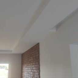 Białe wnętrze z dekoracyjnym sufitem podwieszanym i ścianą z cegły. Widoczne okno z roletami i nowoczesna lampa. Minimalistyczny design.