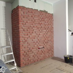 Ściana z cegły dekoracyjnej w trakcie montażu, widoczne puszki elektryczne, płyta gipsowa na górze ściany, drabina i zabezpieczona podłoga.