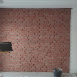 Ściana z cegły dekoracyjnej w jasnym wnętrzu, oświetlona lampami budowlanymi. Widoczny fragment okna i mebli kuchennych. Wykończenie z elementami dekoracyjnymi.