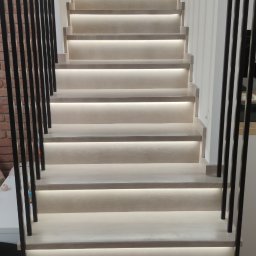 Nowoczesne schody z podświetleniem LED, czarne metalowe barierki, jasne drewno. Ujęcie od dołu, podkreślające minimalistyczny design i subtelne oświetlenie.