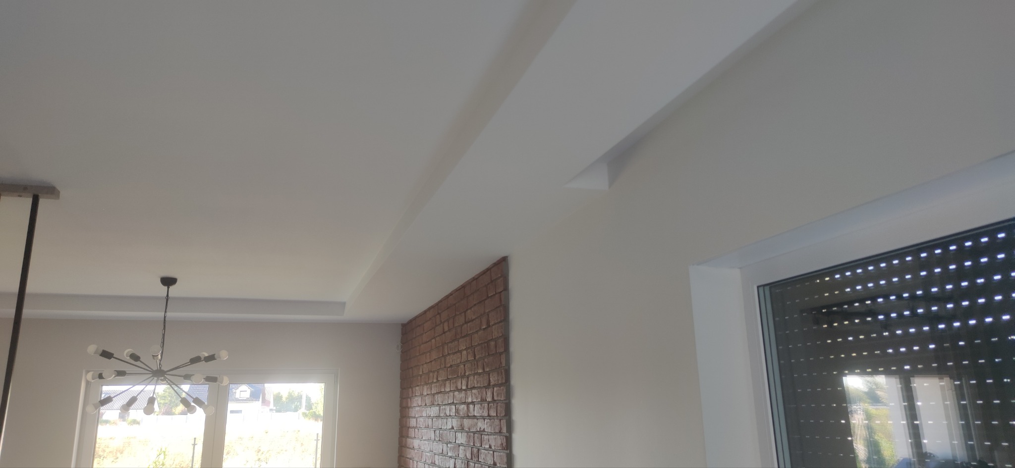 Białe wnętrze z dekoracyjnym sufitem podwieszanym i ścianą z cegły. Widoczne okno z roletami i nowoczesna lampa. Minimalistyczny design.