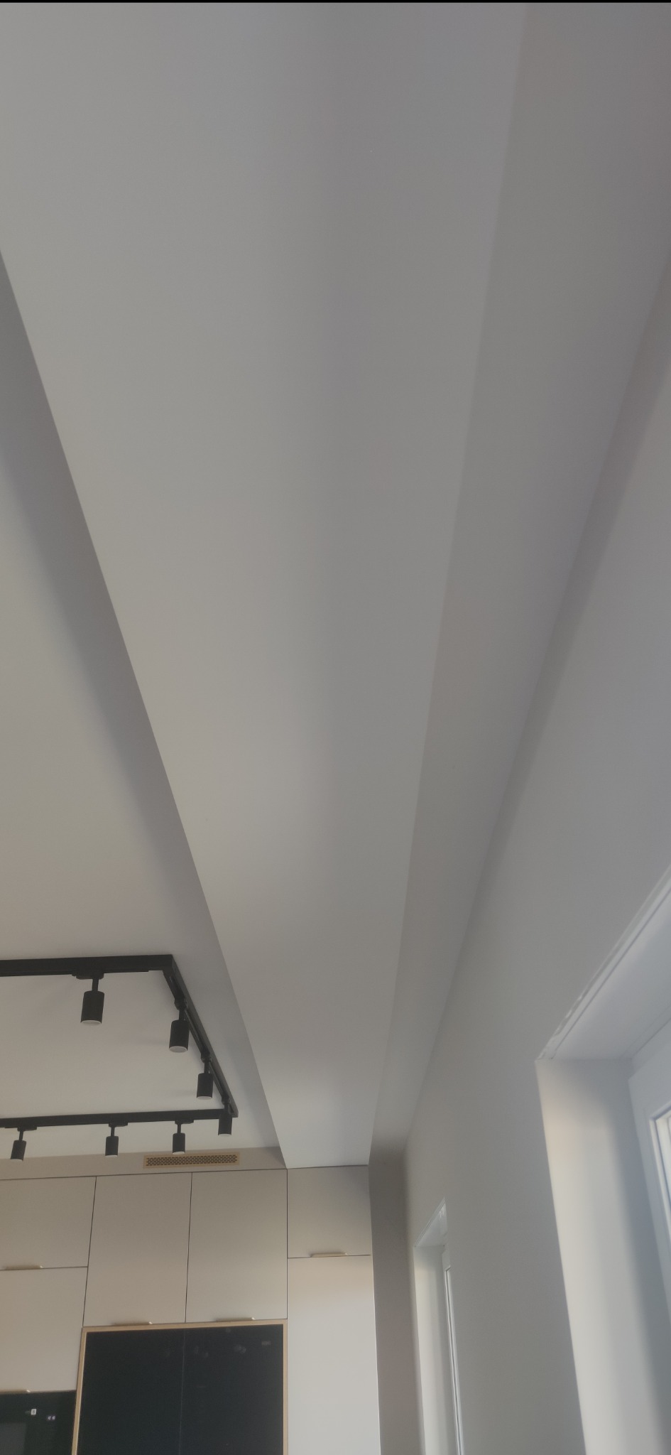Biały sufit z wbudowanym oświetleniem szynowym i subtelnymi wnękami, widok z dołu, fragment zabudowy kuchennej w tle, minimalistyczny design.