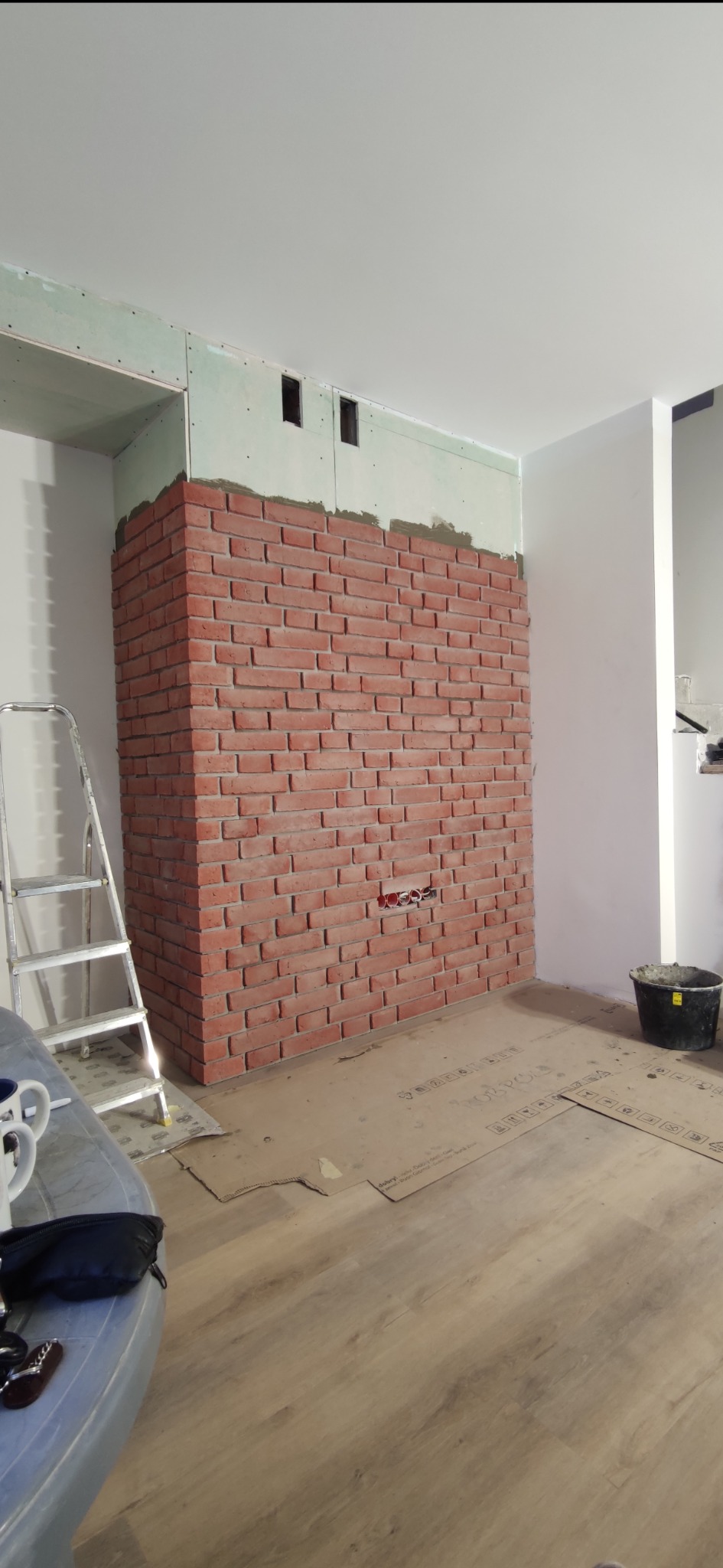 Ściana z cegły dekoracyjnej w trakcie montażu, widoczne puszki elektryczne, płyta gipsowa na górze ściany, drabina i zabezpieczona podłoga.