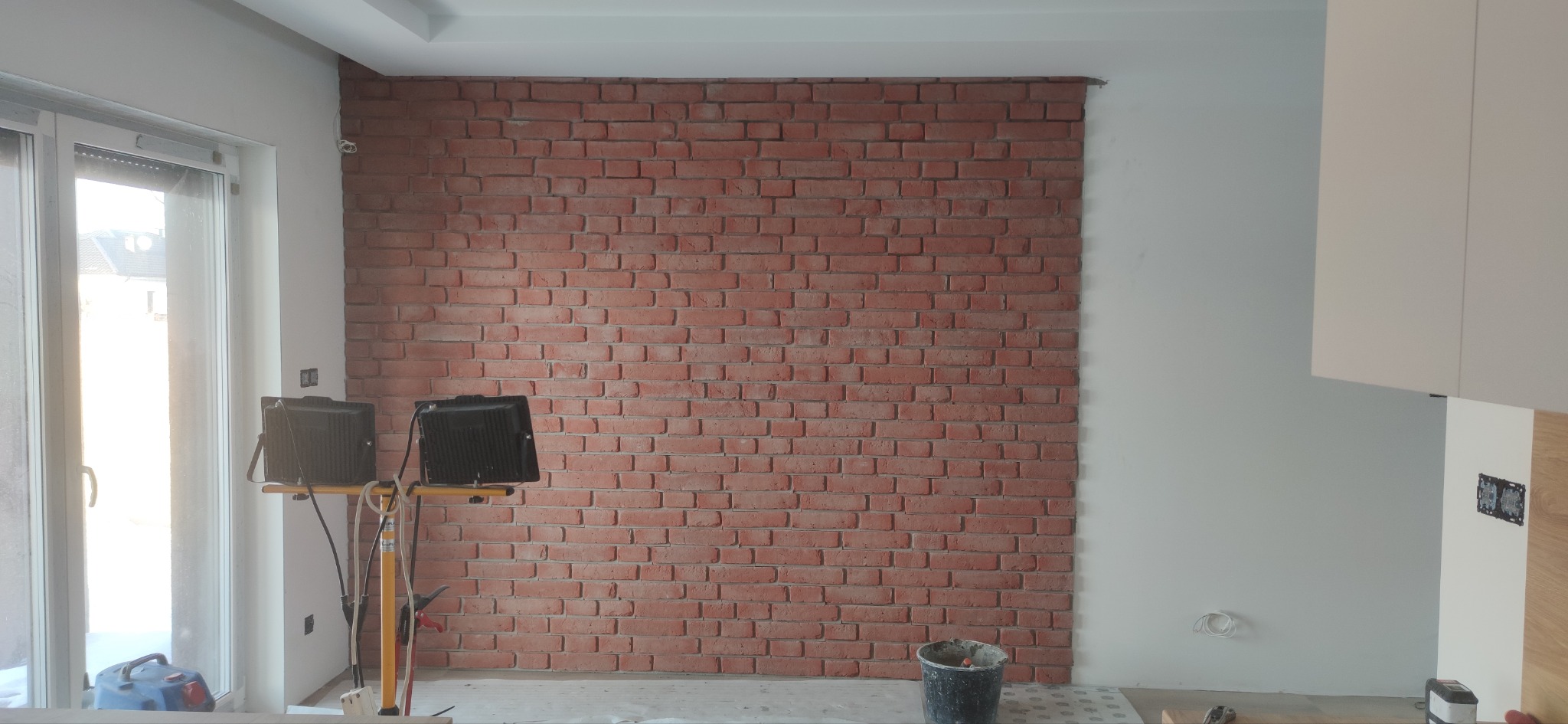 Ściana z cegły dekoracyjnej w jasnym wnętrzu, oświetlona lampami budowlanymi. Widoczny fragment okna i mebli kuchennych. Wykończenie z elementami dekoracyjnymi.