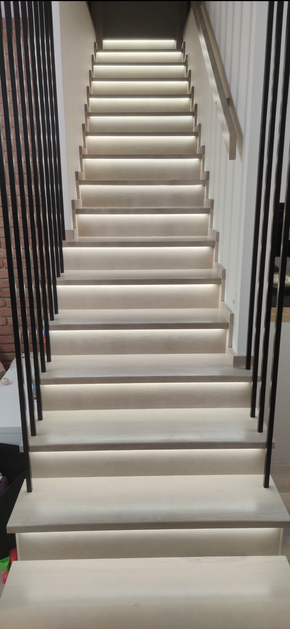 Nowoczesne schody z podświetleniem LED, czarne metalowe barierki, jasne drewno. Ujęcie od dołu, podkreślające minimalistyczny design i subtelne oświetlenie.