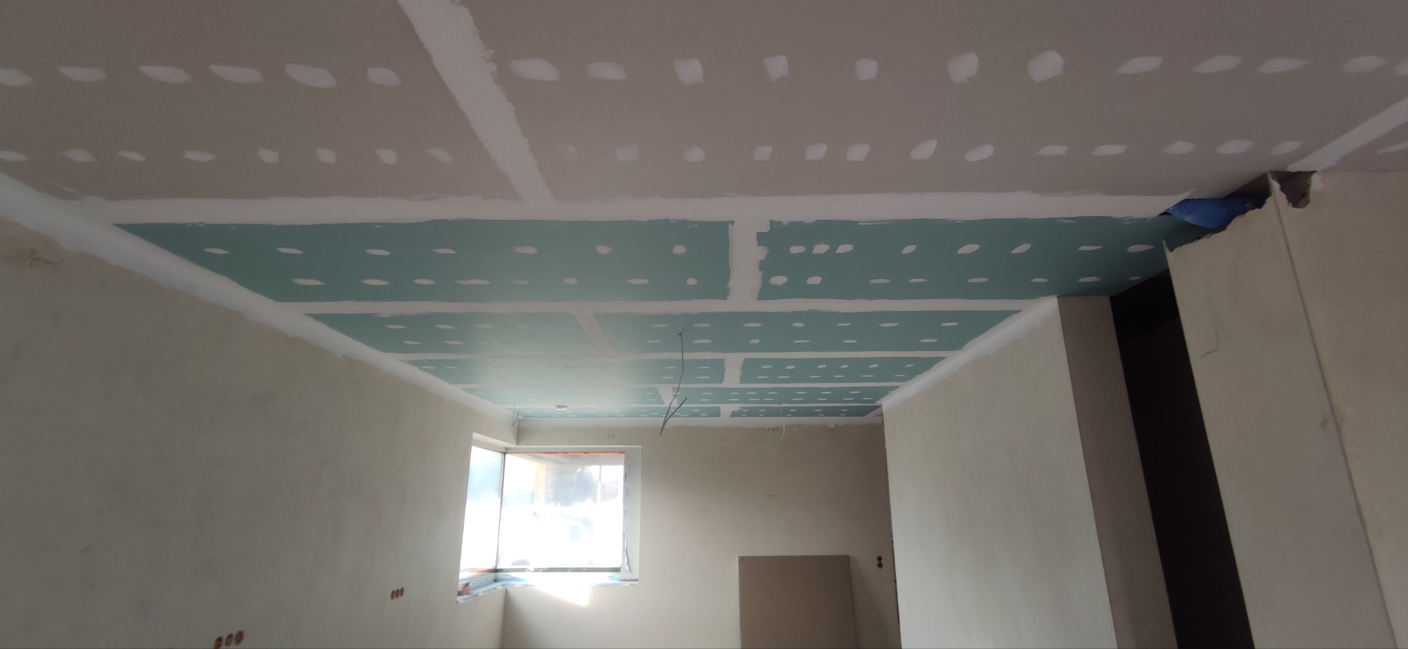 Sufit z płyt gipsowo-kartonowych w trakcie montażu, widoczne łączenia i otwory, surowe ściany, okno w tle. Etap prac wykończeniowych.