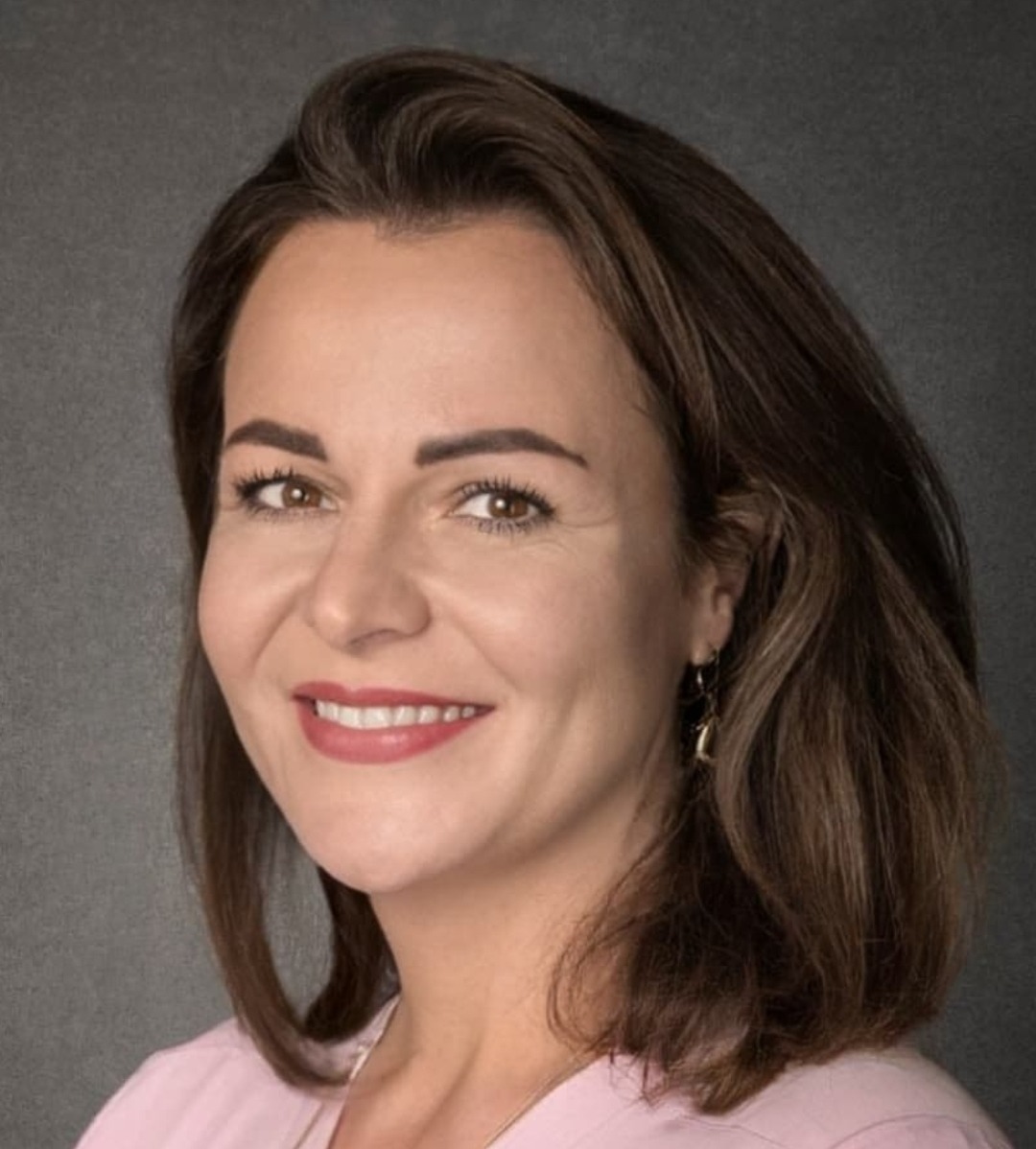 Portret uśmiechniętej brunetki w różowej bluzce, z delikatnym makijażem i złotymi kolczykami na tle szarego tła. Ujęcie od przodu, z lekkim oświetleniem.