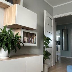 KZ-Project Kamil Ziobroń - Nowoczesny salon z białymi meblami modułowymi, roślinami doniczkowymi i otwartym przejściem do kolejnego pomieszczenia. Minimalistyczny design i jasne wnętrze.