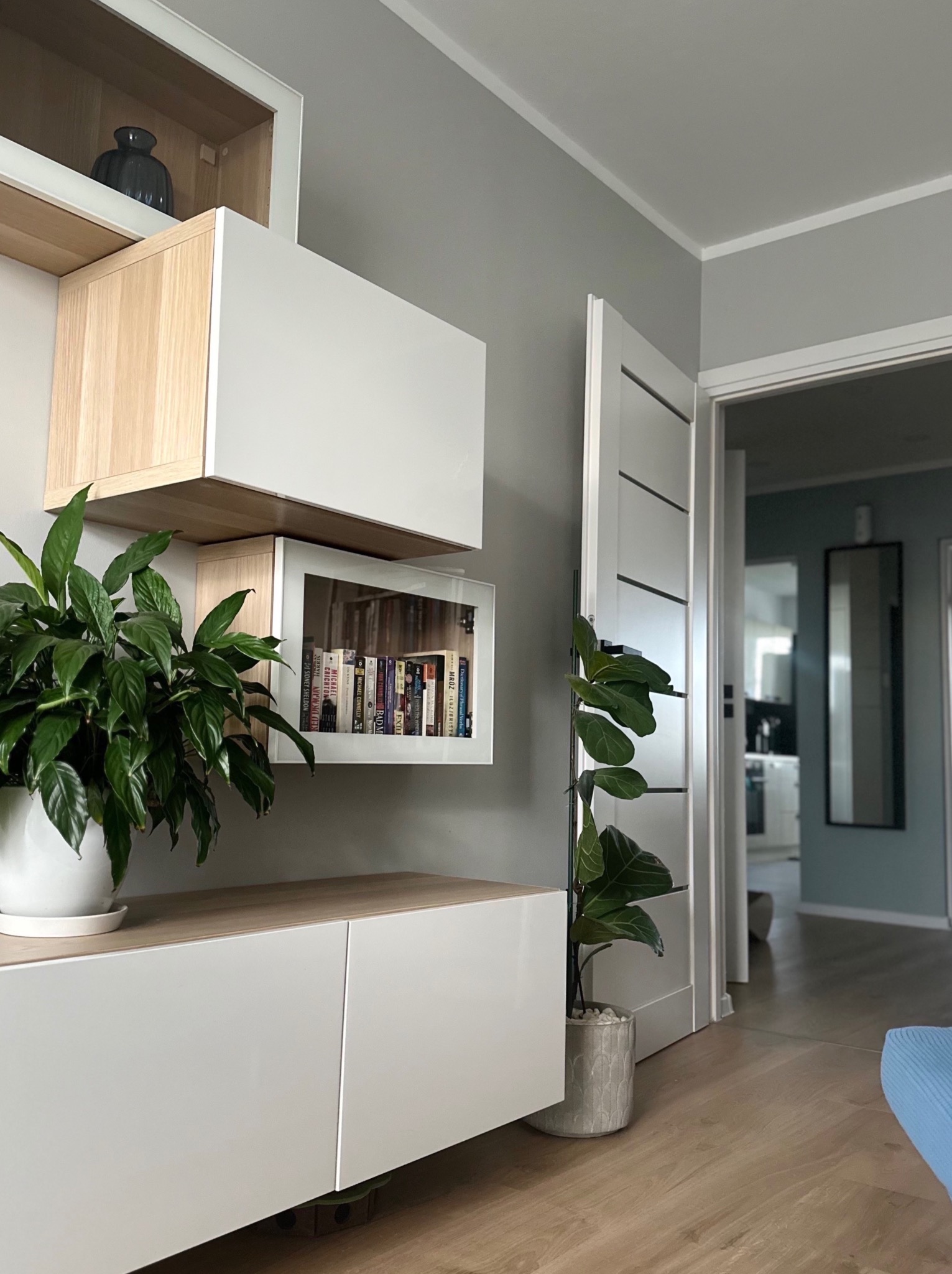 Nowoczesny salon z białymi meblami modułowymi, roślinami doniczkowymi i otwartym przejściem do kolejnego pomieszczenia. Minimalistyczny design i jasne wnętrze.