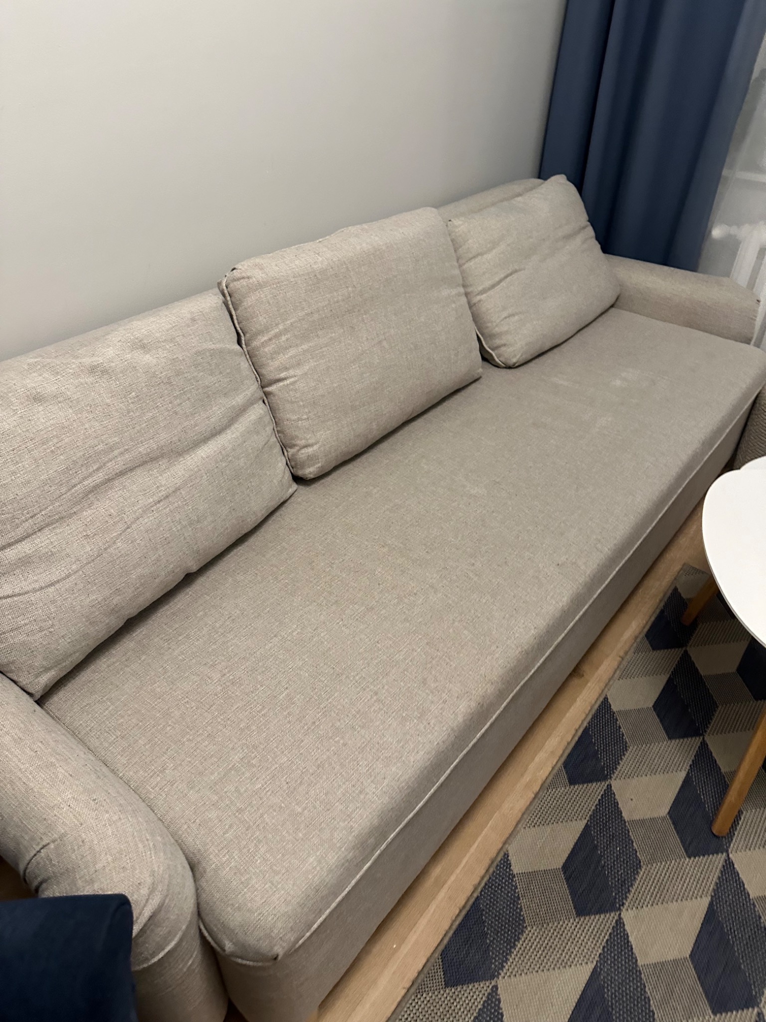 Jasna, trzyosobowa kanapa z poduszkami na tle ściany i wzorzystego dywanu. Widok z góry, częściowo widoczny stolik. Usługa prania kanap w Tarnowie.