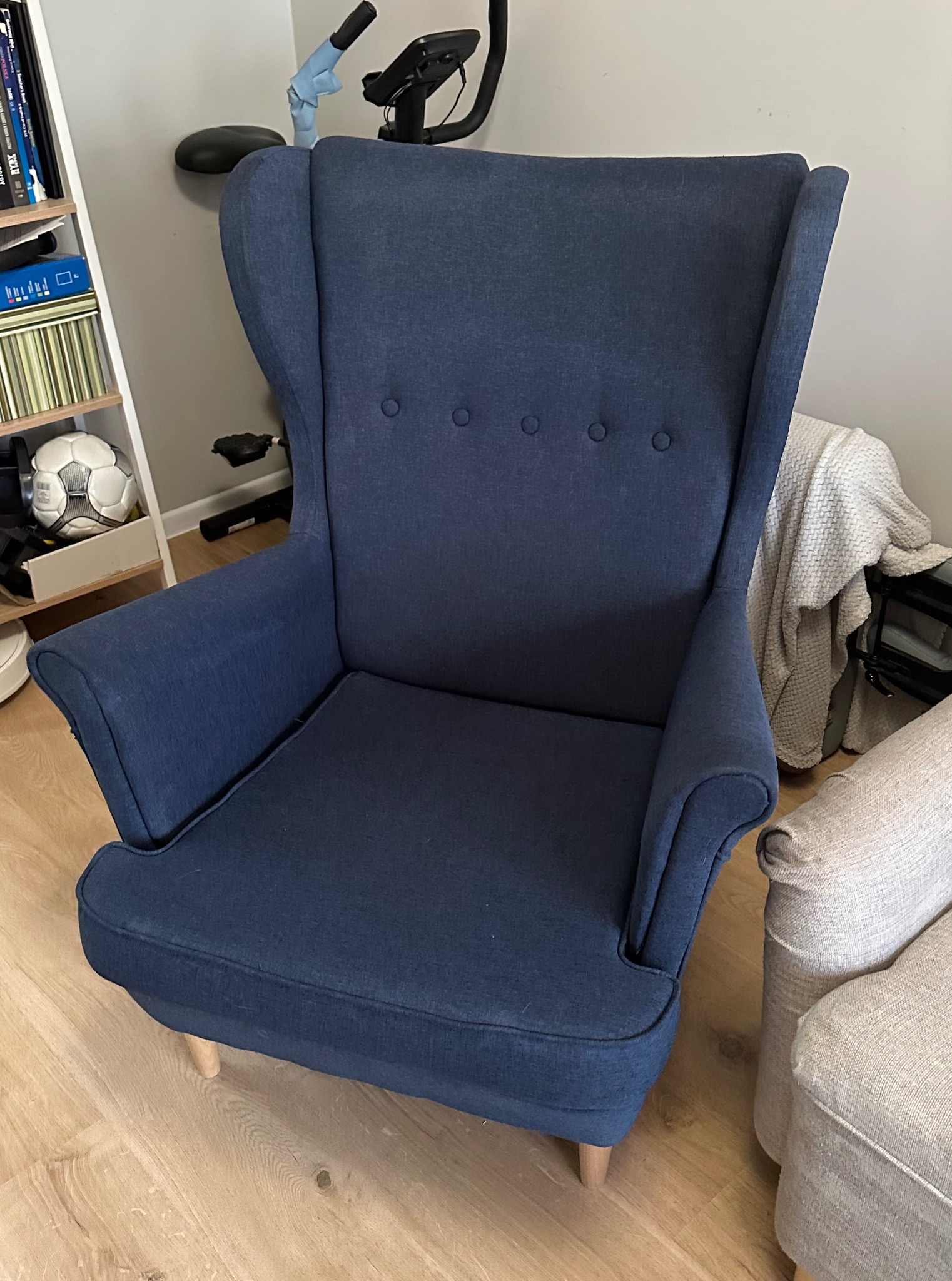 Granatowy fotel typu uszak z guzikami, ustawiony na parkiecie, obok jasnego fotela i regału z książkami. W tle widoczny rower stacjonarny.