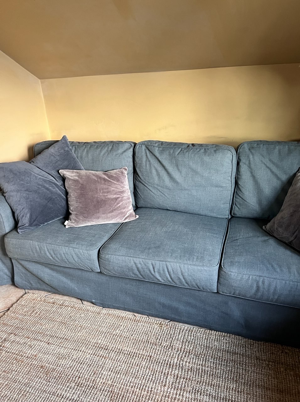 Szara sofa z poduszkami w odcieniach szarości i różu, na tle żółtej ściany i beżowego dywanu. Widok z bliska, wnętrze domu.