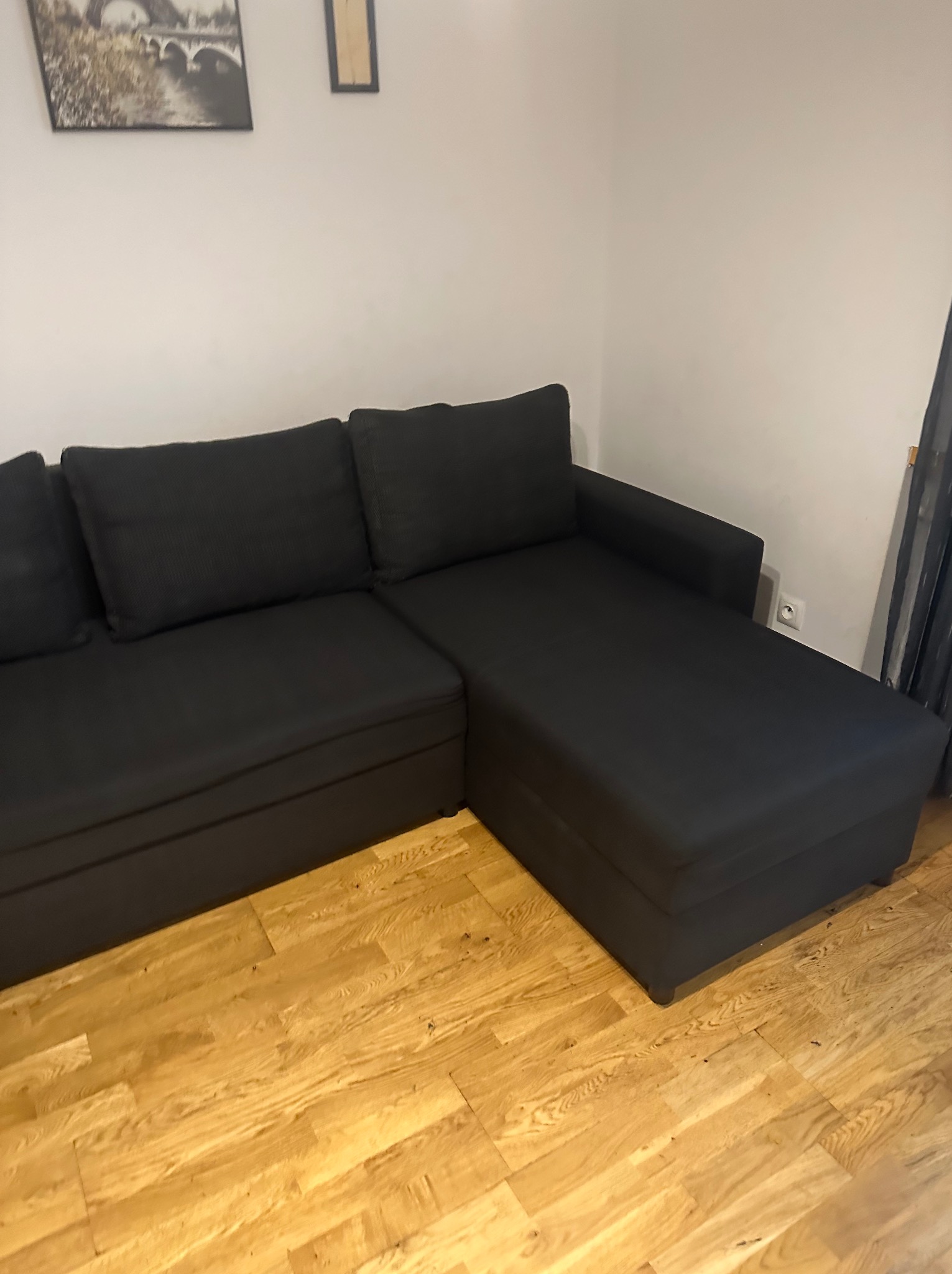 Ciemnoszara, rozłożysta kanapa modułowa na parkiecie, z widocznym gniazdkiem elektrycznym na ścianie i dekoracją ścienną w tle.