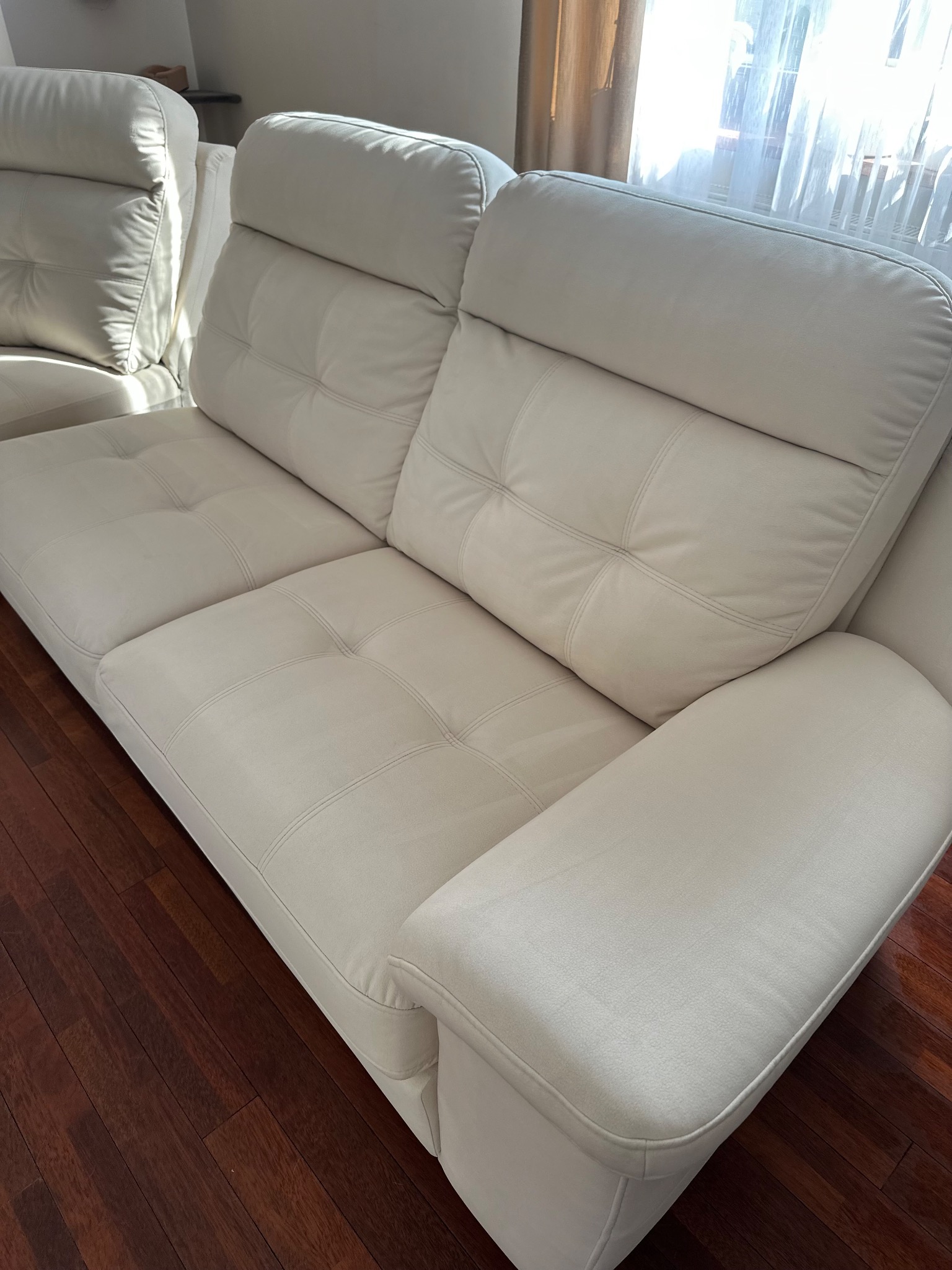 Jasna, skórzana kanapa z pikowanymi siedziskami stoi na ciemnej, drewnianej podłodze. W tle widoczne okno z zasłonami. Detal wnętrza mieszkalnego.