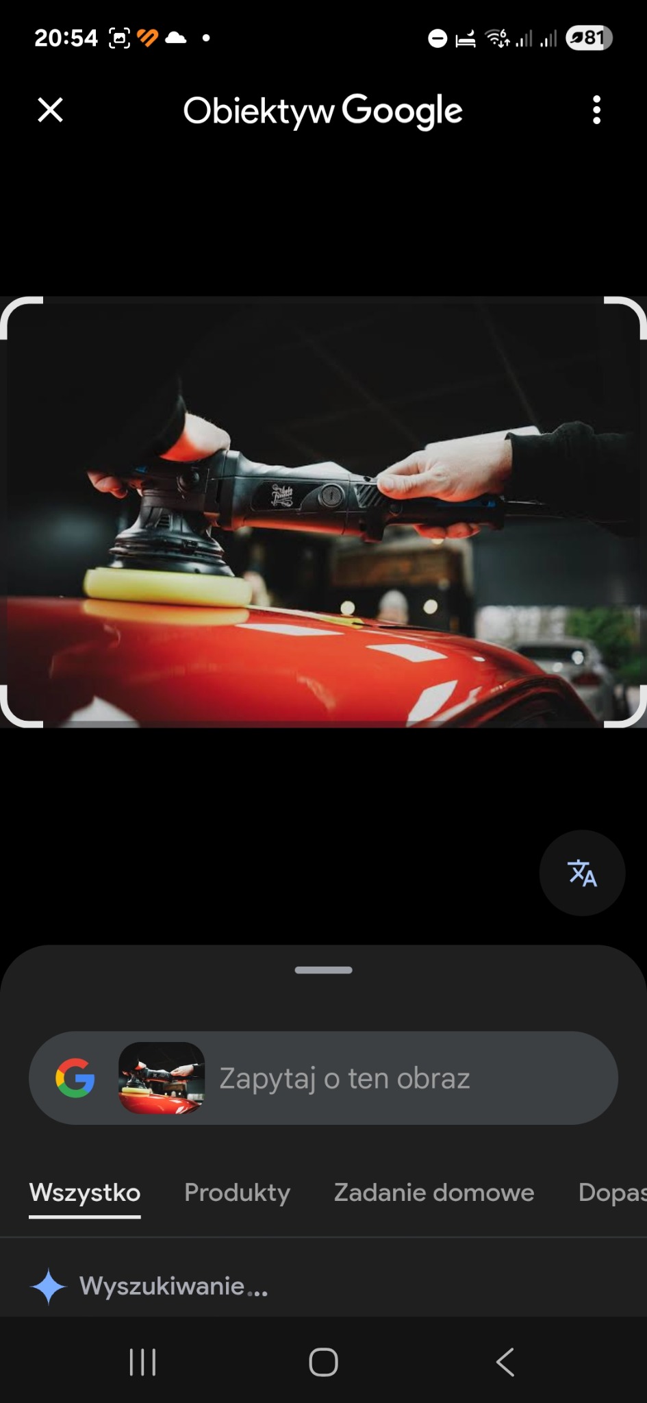 Polerowanie czerwonego lakieru samochodu za pomocą polerki elektrycznej z żółtą gąbką. Dłoń w rękawicy trzyma urządzenie. W tle zarys innego pojazdu.