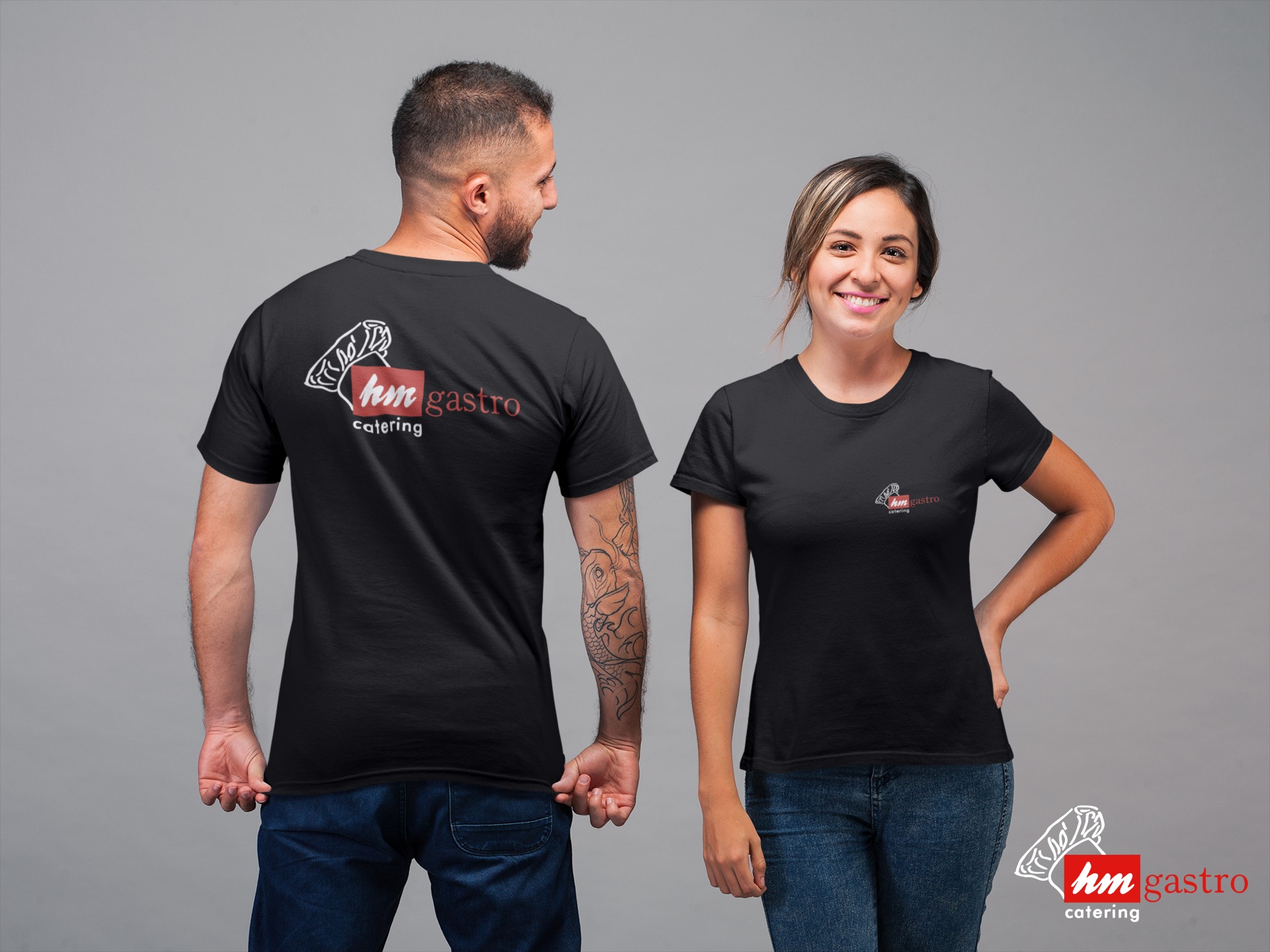 Dwie osoby prezentują czarne koszulki z nadrukiem 'hm gastro catering', mężczyzna z tatuażem na ramieniu, uśmiechnięta kobieta.