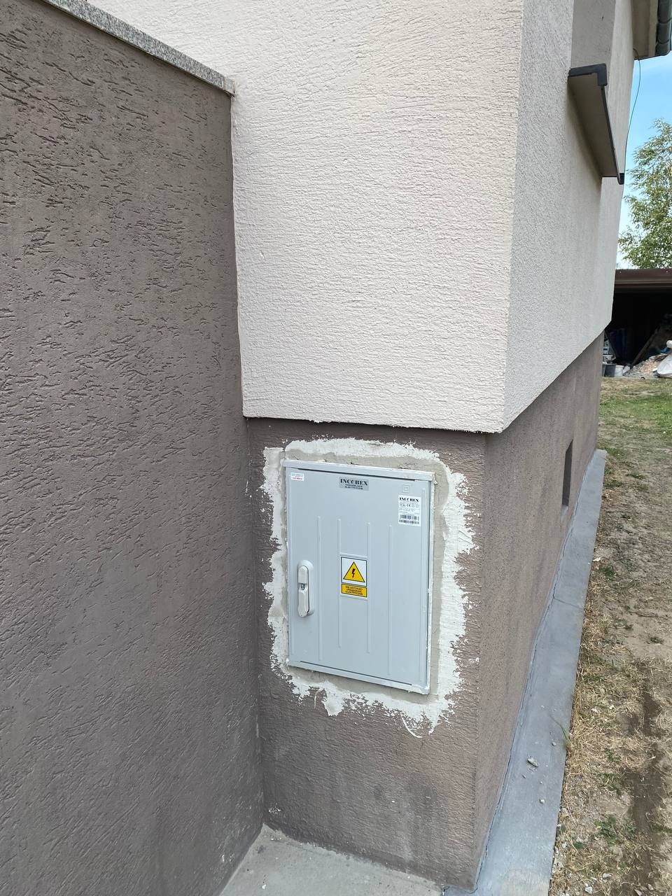 Szara skrzynka elektryczna IncoRex z symbolem ostrzegawczym na elewacji budynku, świeżo osadzona i uszczelniona zaprawą. Widoczny róg budynku i fragment zieleni.